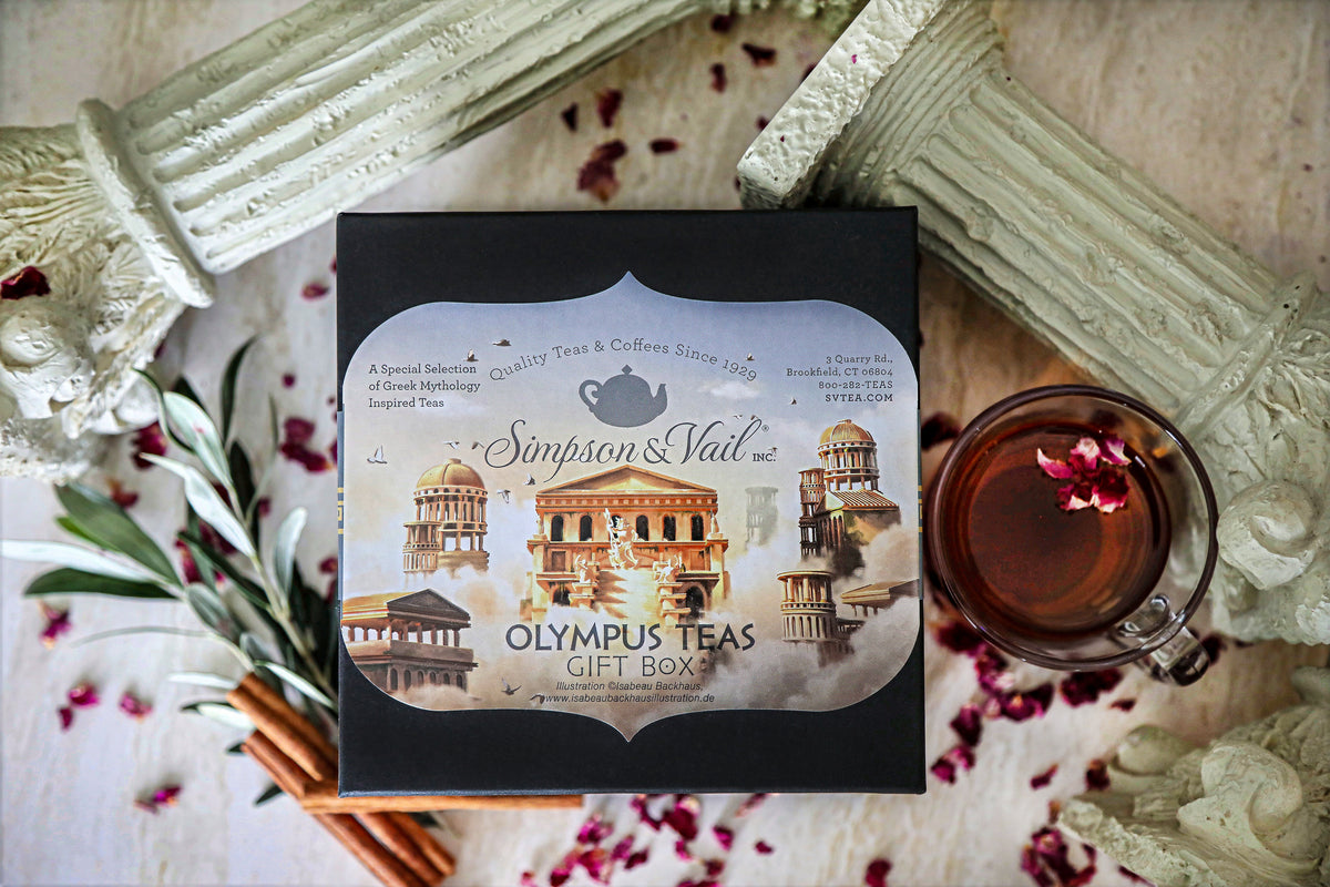 Simpson & Vail Olympus Teas Gift Box in black packaging, styled in a flat-lay with a glass of tea, cinnamon sticks, rose petals, and classical column props on a light wooden surface.